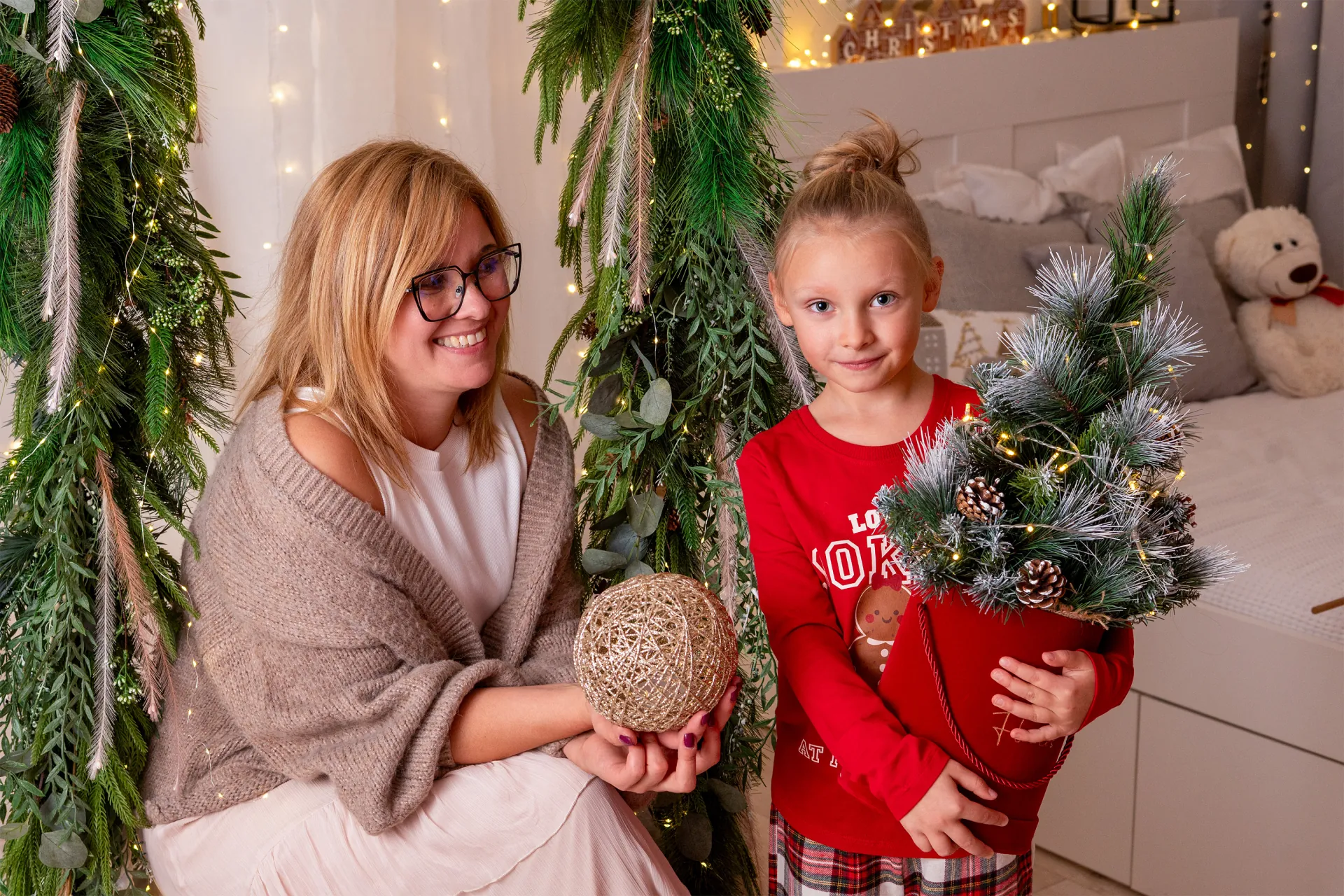 mama z wiklinową kulą i córka z małą ośnieżoną choinką w czerwonej donicy, za nimi girlanda i łóżko z misiem