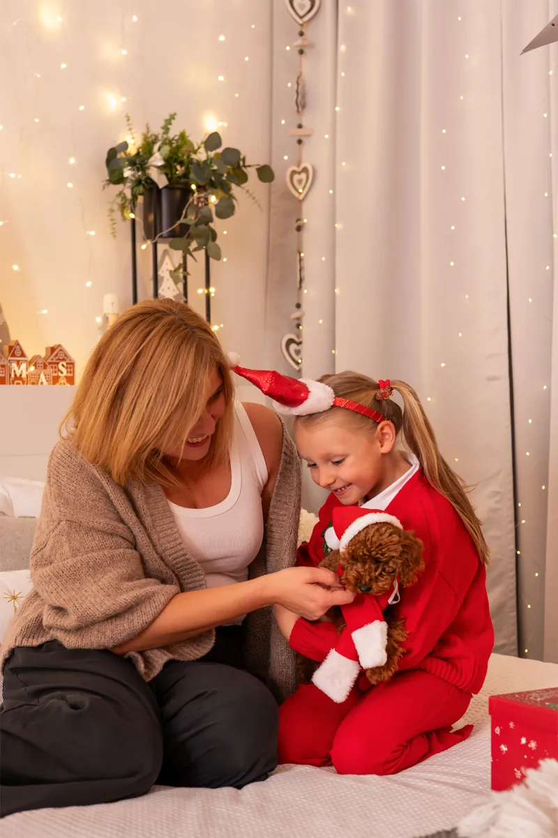 mama i córka w czerwonym stroju bawią się z pudlem w czapce Mikołaja na świątecznym łóżku