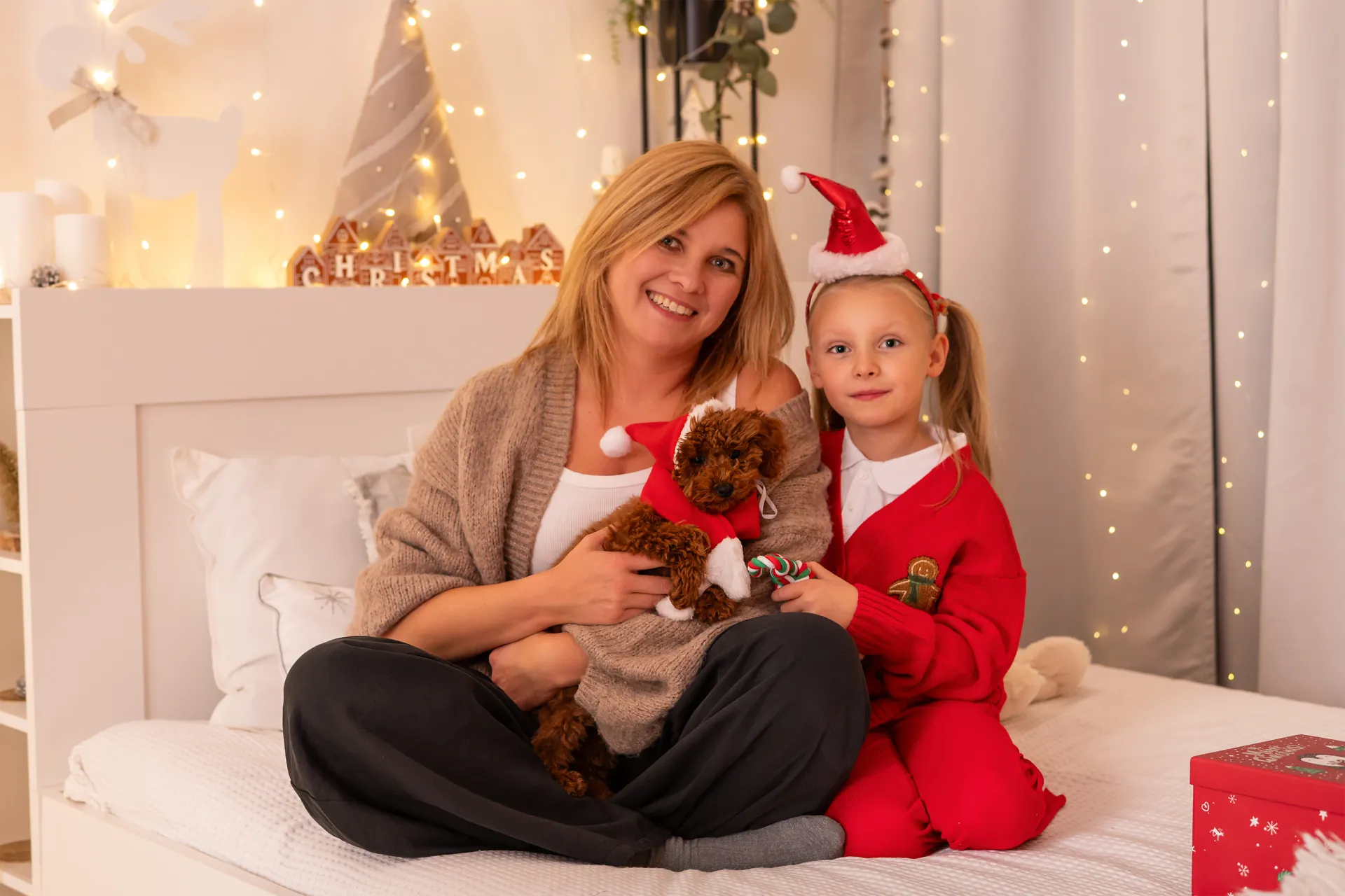 mama z córką i pudlem w czapce Mikołaja na łóżku, za nimi napis Christmas z domków i światełka